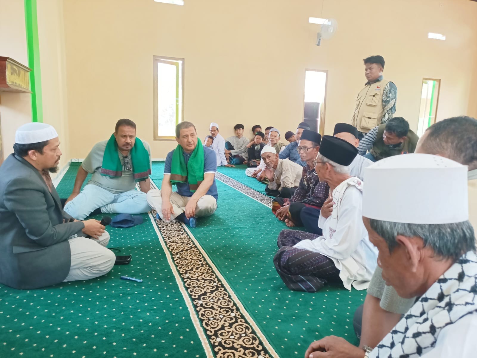 A speaker addresses participants seated in the mosque prayer hall.